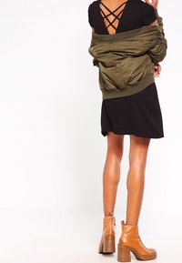 Veste bomber vert olive portée sur une robe noire à dos croisé, associée à des bottines beige à talon empilé.