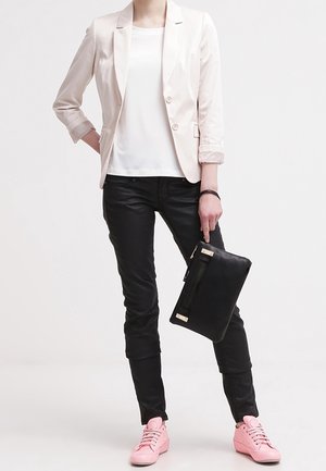 Světlý růžový blejzr přes bílou košili, doplněný černými kalhotami a růžovými teniskami. Drží černé psaníčko se zlatými prvky.