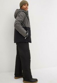 Veste à capuche grise et noire avec une fermeture éclair, en tissu texturé, et poches latérales, associée à un pantalon noir et des bottes noires à semelles marron.