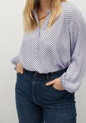 Kobieta o długich blond włosach, ubrana w białą bluzkę w czarne kropki i niebieskie dżinsy, z rękami w kieszeniach i złotym naszyjnikiem.