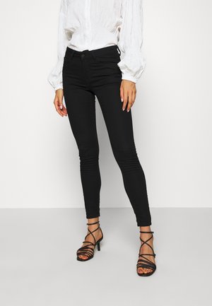 Woman wearing black skinny jeans, white long-sleeve blouse, and black strappy high-heeled sandals standing against white background.