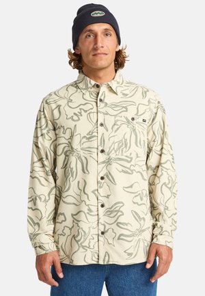 Long-sleeve button-up shirt in cream with green floral pattern, featuring a chest pocket and brown buttons. Worn with a navy beanie.