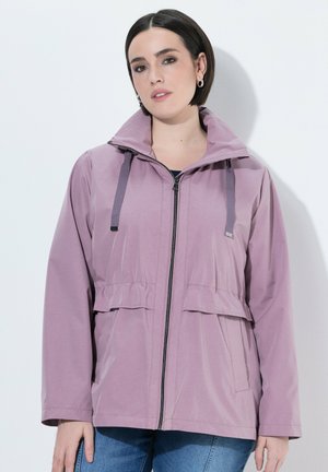 Kvinde med kort mørkt hår iført ringeøreringe, en lys lilla lynlåstrøje med snore og blå jeans mod en hvid baggrund.