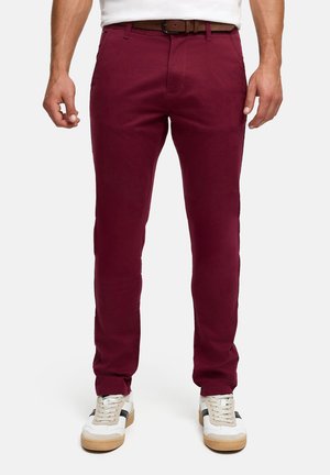 Man draagt slanke bordeauxrode broek met bruine riem en witte sneakers, staand tegen een effen achtergrond.