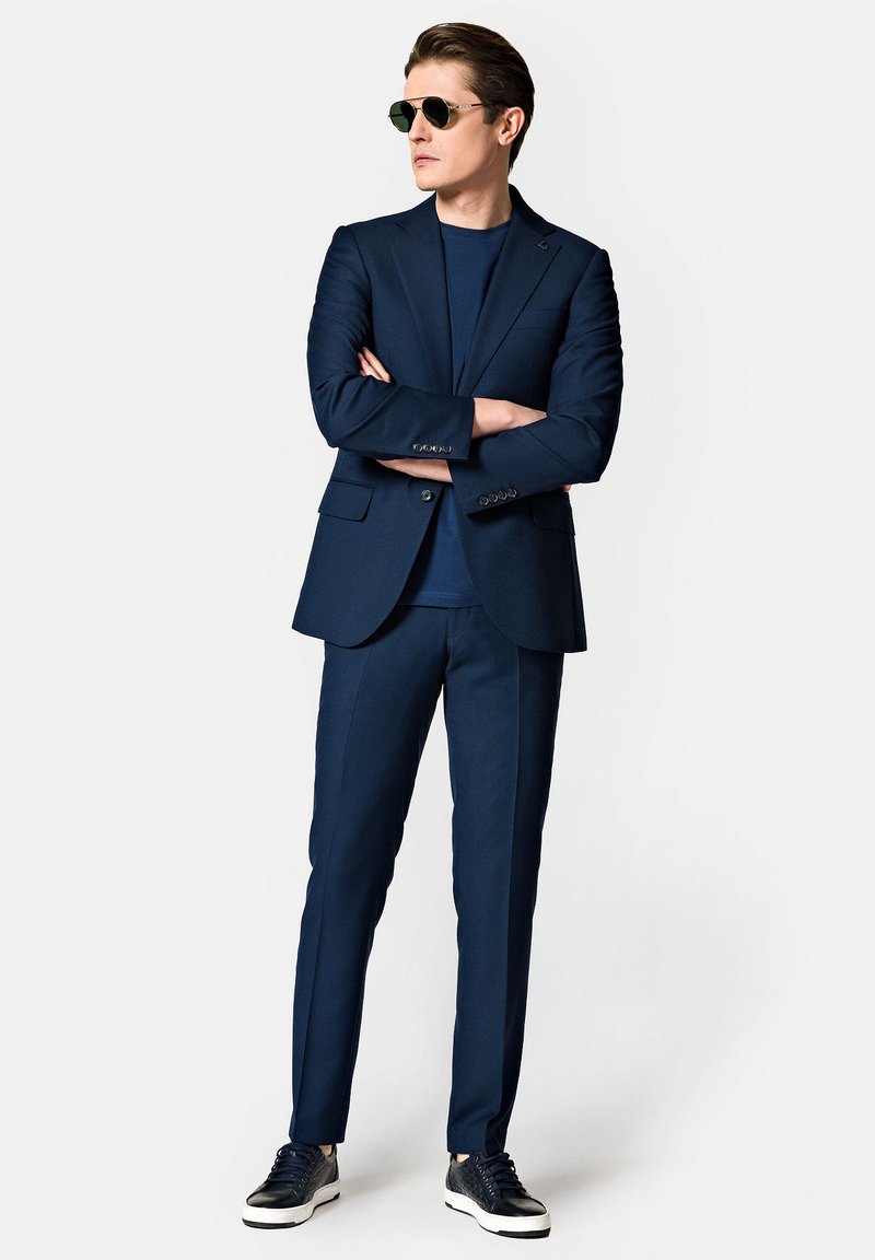Granatowy garnitur z dopasowaną marynarką, pasującymi spodniami slim-fit oraz granatowym t-shirtem. Uzupełniony ciemnymi okularami przeciwsłonecznymi i casualowymi sneakersami.