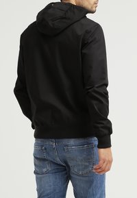 Czarna kurtka z kapturem o gładkim, matowym wykończeniu, z elastycznymi mankietami i luźnym krojem. Noszona z niebieskimi dżinsami z widocznym przeszyciem.