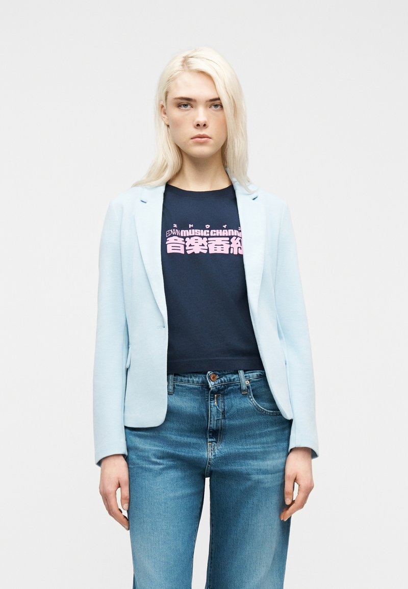 Νεαρή γυναίκα με πλατινένια ξανθά μαλλιά, φορώντας ανοιχτό γαλάζιο σακάκι, μπλε μπλούζα με ροζ γράμματα και μπλε τζιν, στέκεται μπροστά σε απλό φόντο.