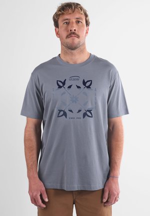 Homme avec des cheveux courts et bouclés et une moustache, portant un t-shirt graphique bleu clair et un pantalon marron sur un fond blanc uni.