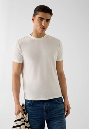 Jeune homme aux cheveux foncés portant un t-shirt en maille texturée blanche à manches courtes et un jean bleu, tenant un vêtement rayé dans sa main gauche.