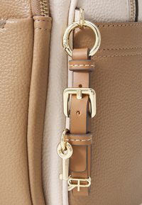 Curea din piele bej cu accesorii aurii, având un sistem de prindere și detalii cu un breloc. Textură netedă, secțiune în contrast de culoare crem.