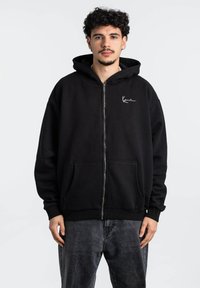 Sudadera con cremallera negra hecha de una mezcla de algodón, que cuenta con un bolsillo frontal y un bordado de logo blanco en el pecho. Textura suave y ajuste relajado.
