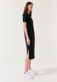 Femme debout de profil portant une robe midi noire à manches courtes avec une fente latérale et des baskets blanches sur un fond uni.