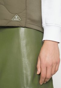 Brązowa pikowana kamizelka z logo Barbour, zestawiona z gładką zieloną skórzaną spódnicą, prezentująca kontrastujące tekstury i kolory.