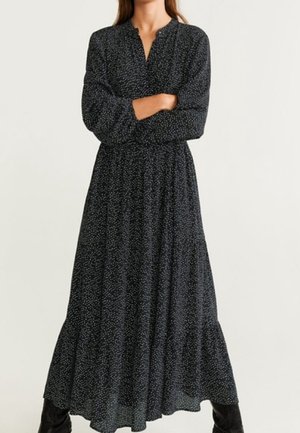 Frau trägt ein langes schwarzes Kleid mit kleinen weißen Punkten, langen Ärmeln und geraffter Taille, steht mit verschränkten Armen.