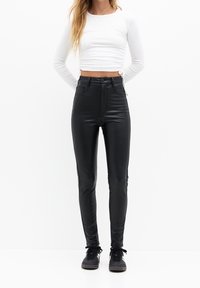 Pantalons noirs en simili cuir taille haute avec une coupe élégante, dotés de poches avant, d'une fermeture éclair et d'un bouton, associés à un haut à manches longues blanc.
