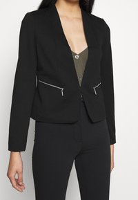 Blazer noir à manches longues, avec des revers nets et deux poches zippées à l'avant. Porté par-dessus un haut boutonné olive. Coupe ajustée.