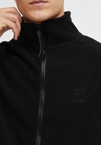 Sort fleece zip-up jakke med høj krave, der har et broderet logo, glat tekstur og kontrastfarvet lynlåsdetalje.