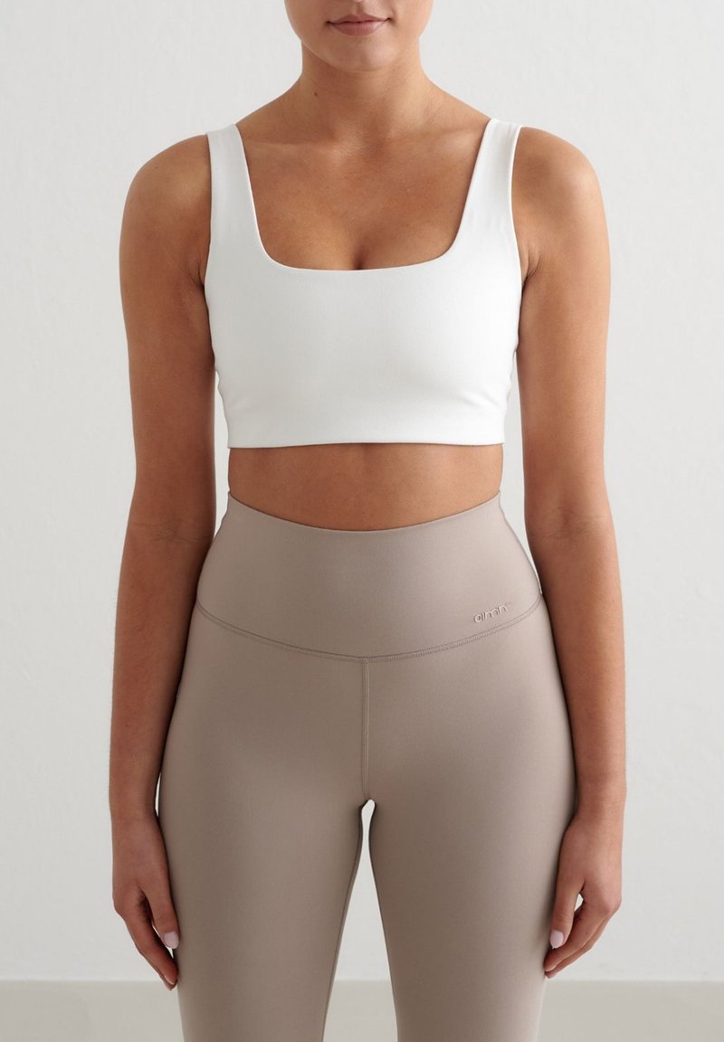 Γυναίκα που φοράει λευκό crop top με τετράγωνο ντεκολτέ και μπεζ κολάν με ψηλή μέση, στέκεται μπροστά σε απλό φωτεινό φόντο.