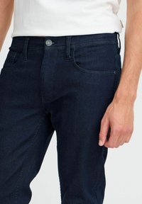 Mörkblå denimjeans med synlig sömnad, en knapp framtill och fem fickor. Tyget verkar slätt med en lätt glans.