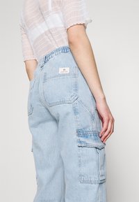 Osoba v světle modrých vysokých cargo džínách s elastickým pasem a průhledném bílém krátkém topu, stojící na jednobarevném pozadí.