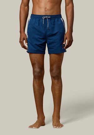 Mannelijk model draagt blauwe zwemshorts met witte zijstrepen en een witte trekkoord, staat blootsvoets tegen een effen achtergrond.