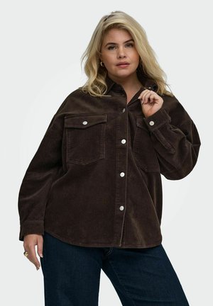 Blonde Frau trägt eine dunkelbraune Cordjacke mit Fronttaschen und dunkelblaue Jeans, posiert mit einer Hand nahe am Kragen.