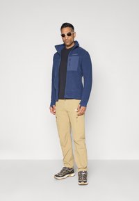 Giacca in pile blu navy con zip frontale, tessuto strutturato, tasca sul petto, abbinata a pantaloni beige e scarpe da hiking grigie. Occhiali da sole bianchi.