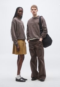 Dos modelos están uno al lado del otro. Uno lleva una sudadera marrón y una falda a cuadros amarilla; el otro tiene una sudadera marrón a juego con pantalones.