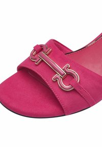 Růžové semišové sandály na podpatku s širokým páskem a velkou dekorativní kovovou sponou s lesklým povrchem. Podešev je černá s mírným zakřivením.