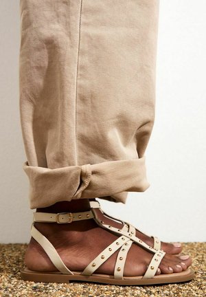 Beige leren sandalen met gespikkelde riemen, verstelbare enkelgesp en platte zool, gecombineerd met opgerolde lichtbeige broeken.