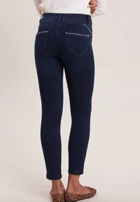 Jeans skinny blu scuro con una texture liscia, dotati di tasche posteriori e dettagli con zip argentate sui lati. Orlo arrotolato alle caviglie.