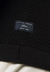 Schwarzer Strickstoff mit einem strukturierten Muster; verfügt über ein dunkelblaues Etikett mit der Aufschrift "s.Oliver" und "Clothing Company Since 1969" auf einem schwarzen Besatz.