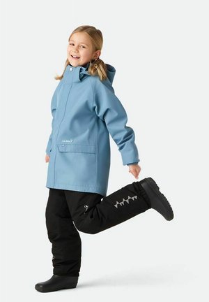 Smilende barn iført en blå regnjakke og sorte vandtætte bukser, løfter det ene ben, mens det står op mod en ensfarvet baggrund.