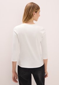 Weißes Langarmshirt aus weichem Baumwollstoff, mit einem runden Ausschnitt und einem leicht abgerundeten Saum. Getragen mit schwarzen Hosen.