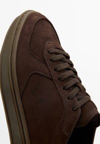 Brun mockasneaker med rund tå, platta snören och strukturerade sidopaneler. Har en tjock gummisula och sömmar som detaljer.