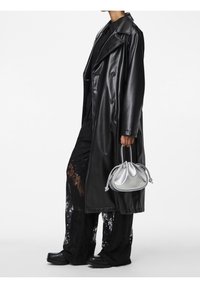 Trench-coat en similicuir noir avec un large col et des fermetures à boutons, associé à un pantalon noir à motifs et un petit sac à main argenté.