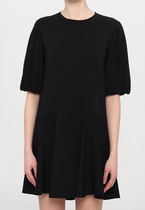 Kobieta ubrana w czarną sukienkę z krótkimi bufiastymi rękawami i lekko rozkloszowanym dołem, stojąca na jednolitym tle.