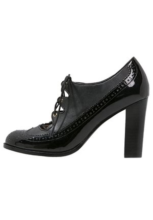 Zwarte leren en lakleren oxford damesschoen met hoge hak, brogue-details en vetersluiting aan de voorkant, zijaanzicht op witte achtergrond.