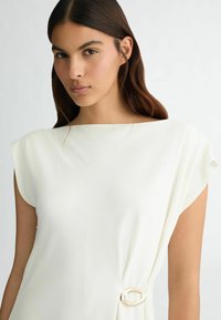 Abito bianco con maniche corte a cappuccio, caratterizzato da un dettaglio con nodo laterale decorato con un accento circolare. Tessuto liscio e strutturato con un design drappeggiato.