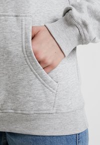 Sudadera gris clara con un bolsillo tipo canguro, puños y dobladillo de canalé. La tela tiene una textura sutil. Jeans de mezclilla son parcialmente visibles.