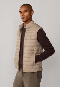  chaleco acolchado beige con cuello alto, cierre de cremallera y bolsillos laterales, llevado sobre un suéter marrón oscuro, combinado con pantalones marrones.