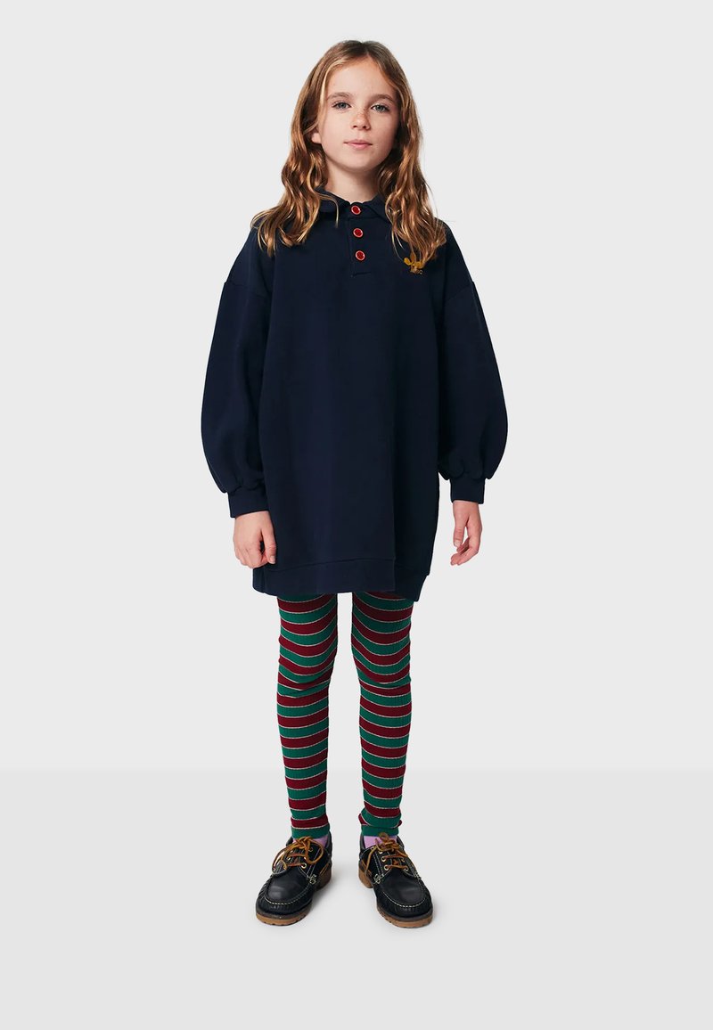 Robe pull oversized bleu marine avec trois boutons rouges au col, associée à des leggings rayés multicolores verts et rouges. Chaussures noires.