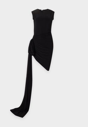 Femme portant une robe noire asymétrique sans manches avec un détail drapé sur un côté et des sandales noires à talons, debout, la main sur la hanche.