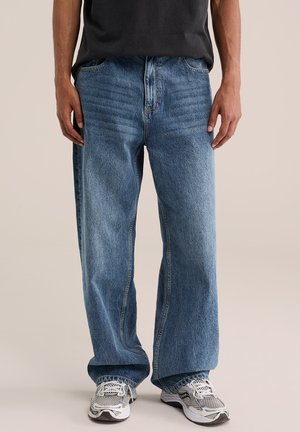 Homme portant un jean bleu ample et des baskets blanches, debout devant un fond uni, les mains détendues le long du corps.