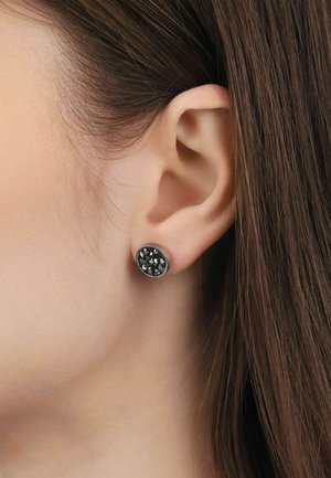 Nahaufnahme eines linken Ohrs mit einem runden schwarzen Stecker-Ohrschmuck mit kleinen reflektierenden silbernen Sprenkeln vor glattem braunem Haar.
