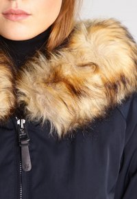 Laivastonsininen takki irrotettavalla keinoturkiskauluksella, jossa on vaaleanruskeita ja tummia raitoja, vetoketjusuljenta ja sileä kankaan rakenne.