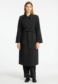 Dolge črne plašč herringbone vzorca z dvojnim zapenjanjem s gumbi, velikim ovratnikom in ujemajočim pasom. Ima široka rokava in teksturirano tkanino.