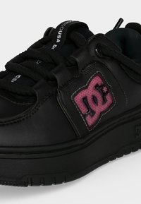 Baskets noires avec semelle épaisse, lacets noirs et logo DC rose sur le côté avec couture blanche sur fond uni.