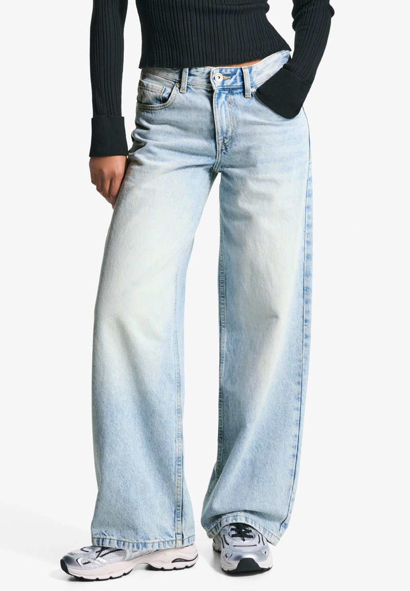 Jean large bleu clair en denim, à taille haute, avec cinq poches et un motif délavé et usé. Portés avec des baskets argentées.