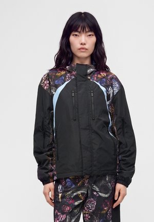 Femme aux longs cheveux noirs porte une veste noire avec des motifs floraux et papillons et un pantalon assorti avec des serpents et des fleurs.